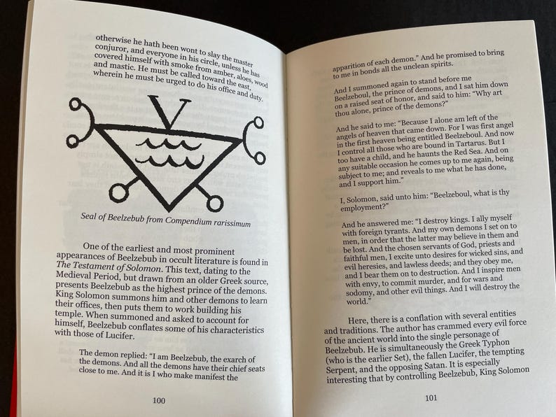May include: An open book with black text on white pages. The left page features a black line drawing of the Seal of Beelzebub, a triangle with a V on top and wavy lines inside. The text discusses the Testament of Solomon and Beelzebub.