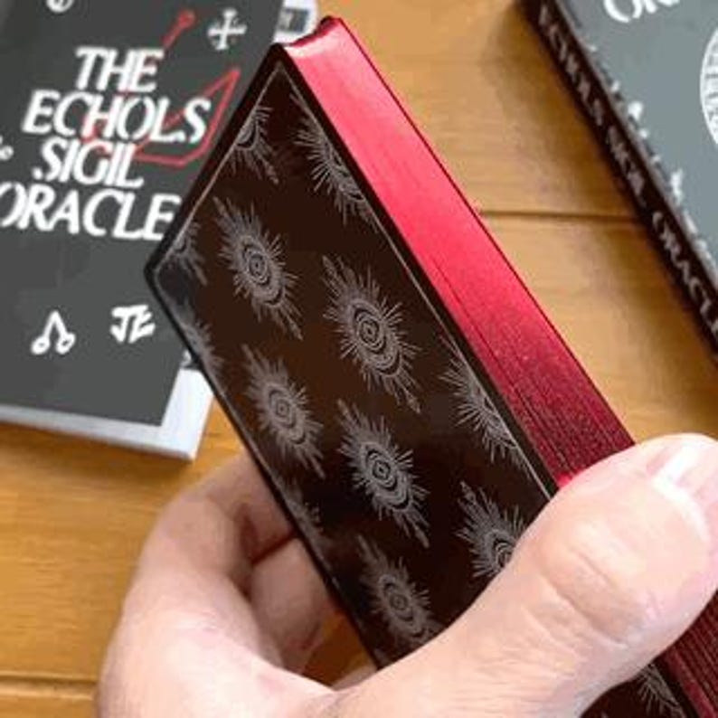 May include: The back of a black deck of oracle cards with a red gilded edge. The cards feature a repeating pattern of a white eye with radiating lines.
