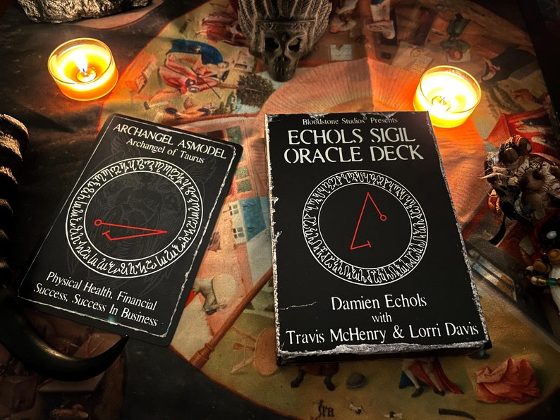 May include: Two black oracle cards with red sigils and white text. The card on the left reads "Archangel Asmodel, Archangel of Taurus, Physical Health, Financial Success, Success in Business". The card on the right reads "Bloodstone Studios Presents, Echols Sigil Oracle Deck, Damien Echols with Travis McHenry & Lorri Davis".