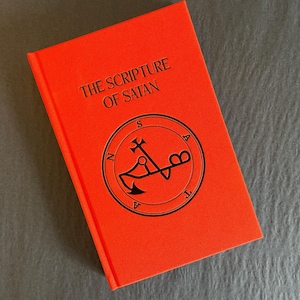 May include: A bright orange hardcover book titled The Scripture of Satan in black lettering. The cover features a circular black emblem with the word Satan spelled out around the perimeter and a central symbol resembling an anchor and cross.