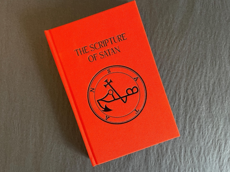 May include: A bright orange hardcover book titled The Scripture of Satan in black lettering. The cover features a circular black emblem with the word Satan spelled out around the perimeter and a central symbol resembling an anchor and cross.