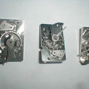 May include: Three silver watch movements with intricate gears and hands are displayed against a white background. The movements are rectangular, showcasing the internal mechanics of a timepiece. The hands point to different times, highlighting the functionality of the watch parts.