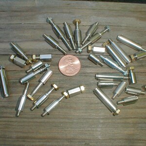 May include: A collection of small, shiny metal fasteners with hexagonal heads and cylindrical bodies, arranged on a wooden surface. A penny is in the center for size comparison. The fasteners vary in length and design, with some having threaded ends.