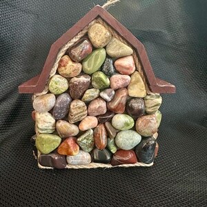 May include: A small wooden house shaped ornament covered in colorful stones and accented with twine.