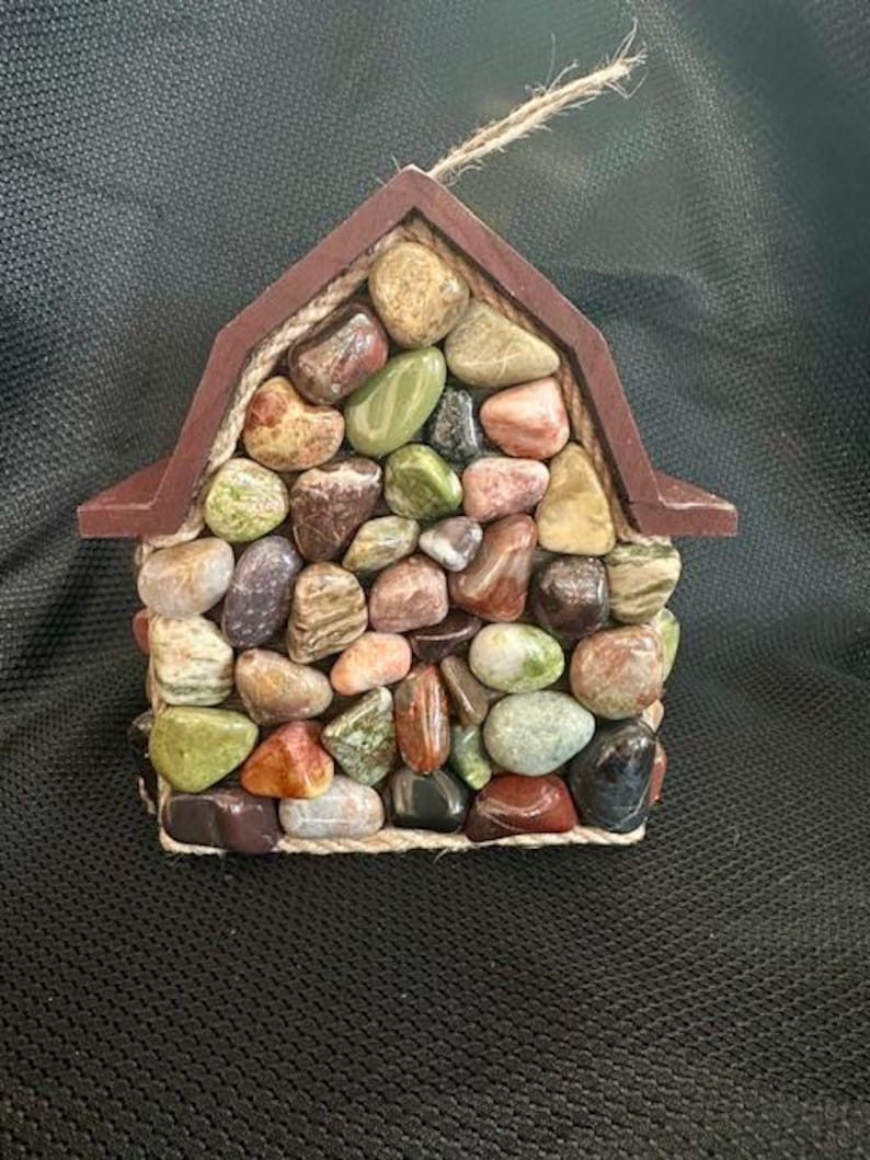 May include: A small wooden house shaped ornament covered in colorful stones and accented with twine.