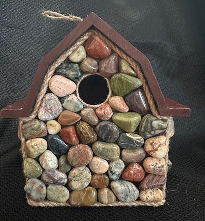May include: A wooden birdhouse with a brown roof and a front made of colorful, smooth stones. The stones are arranged in a mosaic pattern and are held in place with twine.