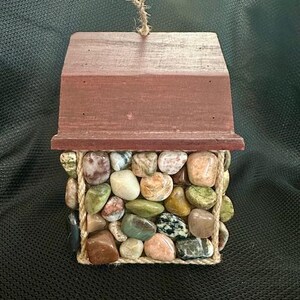 May include: A small wooden house with a red roof and a rope handle. The house is covered in a mosaic of colorful stones.