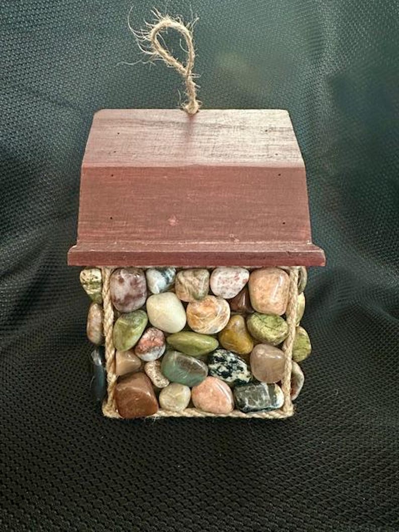 May include: A small wooden house with a red roof and a rope handle. The house is covered in a mosaic of colorful stones.