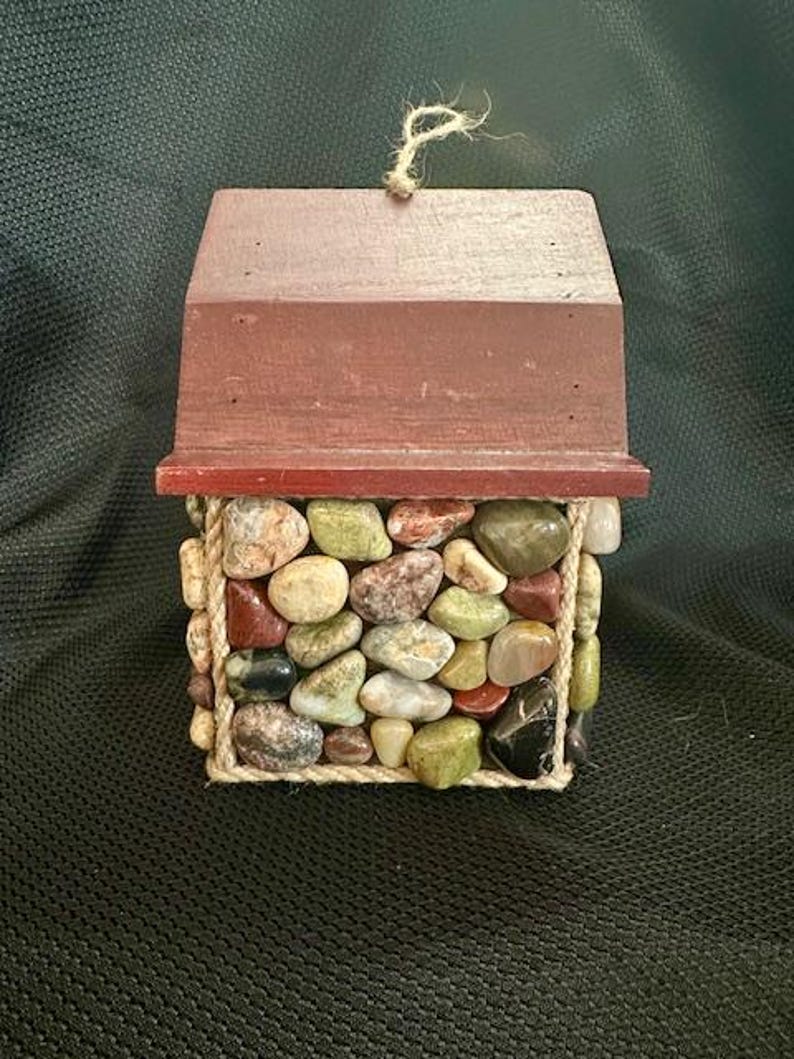 May include: A small wooden house with a red roof, decorated with a variety of colorful stones and wrapped with twine.