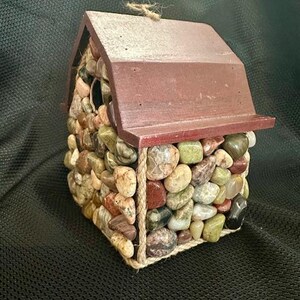 May include: A small wooden birdhouse with a red roof, covered in colorful stones and accented with twine.