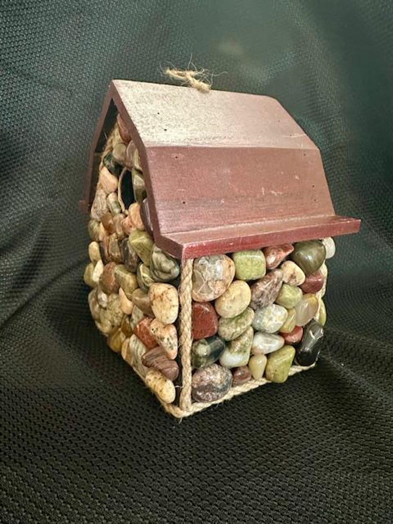 May include: A small wooden birdhouse with a red roof, covered in colorful stones and accented with twine.