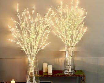 May include: Two sets of white twig branches with warm white LED lights in clear glass vases on a brown wooden table.