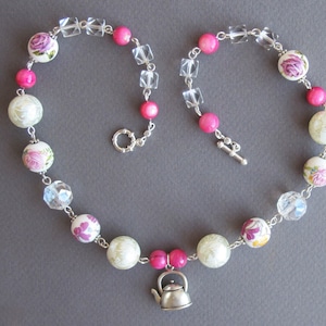 Puede incluir: Un collar de cuentas blancas y rosas con un dije de tetera plateada. Las cuentas tienen diseños florales y detalles de cristal transparente.