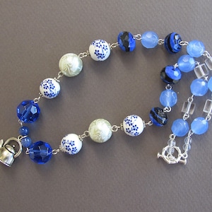 Puede incluir: Un collar de cuentas de cerámica azul y blanco con un dije de tetera de plata. El collar presenta un degradado de cuentas azules, incluyendo cuentas azul claro, azul oscuro y cuentas con patrones en blanco y negro.