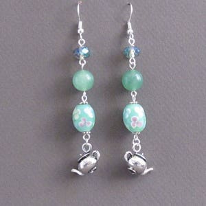 Könnte beinhalten: Ein Paar Ohrhänger mit silberfarbenen Haken. Jeder Ohrring hat eine blaue Kristallperle, eine grüne Perle und eine türkisfarbene ovale Perle mit floralem Design. Ein kleiner silberfarbener Teekannen-Anhänger hängt unten.