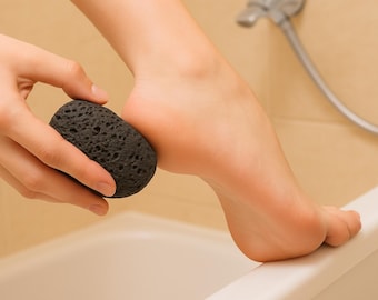 Puede incluir: Una piedra pómez negra y porosa se utiliza en un pie. La piedra, de forma ovalada, se sostiene en una mano. El pie descansa sobre el borde de una bañera blanca. El fondo es de color beige claro.