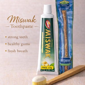 Op de afbeelding: Afbeelding van Dabur Miswak tandpasta, een Miswak-stok en een bamboe tandenborstel. De tube toont de naam "Miswak" en benadrukt voordelen zoals sterke tanden en een frisse adem.