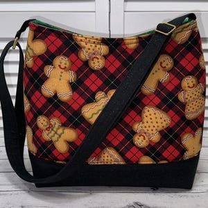 May include: A black and red plaid tote bag with a gingerbread man pattern. The bag has a black adjustable strap and a black bottom.