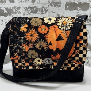 May include: A crossbody bag featuring a flap with a black, orange, and cream print of a jack-o'-lantern, flowers, bats, and a checkered pattern. The bag has a solid black base, a black adjustable strap, and a silver-toned metal twist-lock closure.