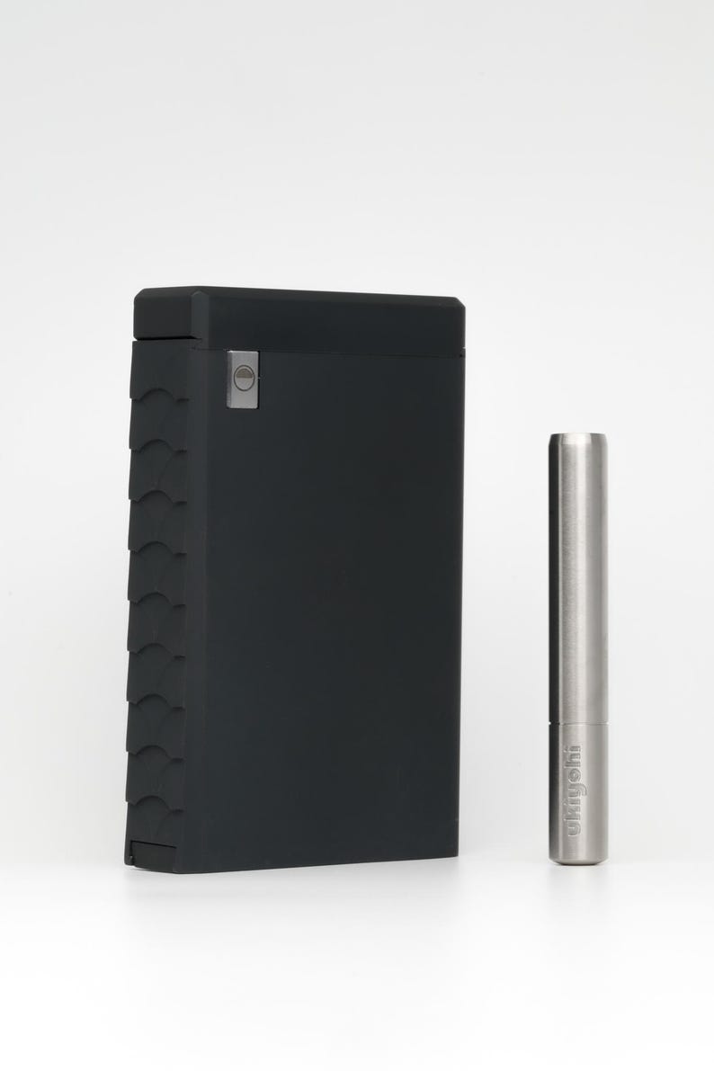 May include: A black, textured, rectangular case with a silver button on the front. Next to it is a silver metal tool with the word "wryeh" engraved on it.