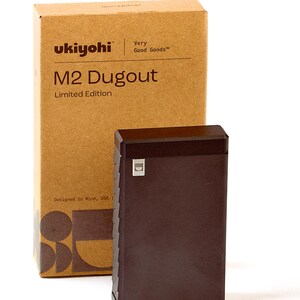 May include: A brown, rectangular box with the text "ukiyohi" and "M2 Dugout Limited Edition" printed on the front. The box is sitting on a white surface next to a brown, rectangular, metal container with a small, silver, rectangular logo on the front.