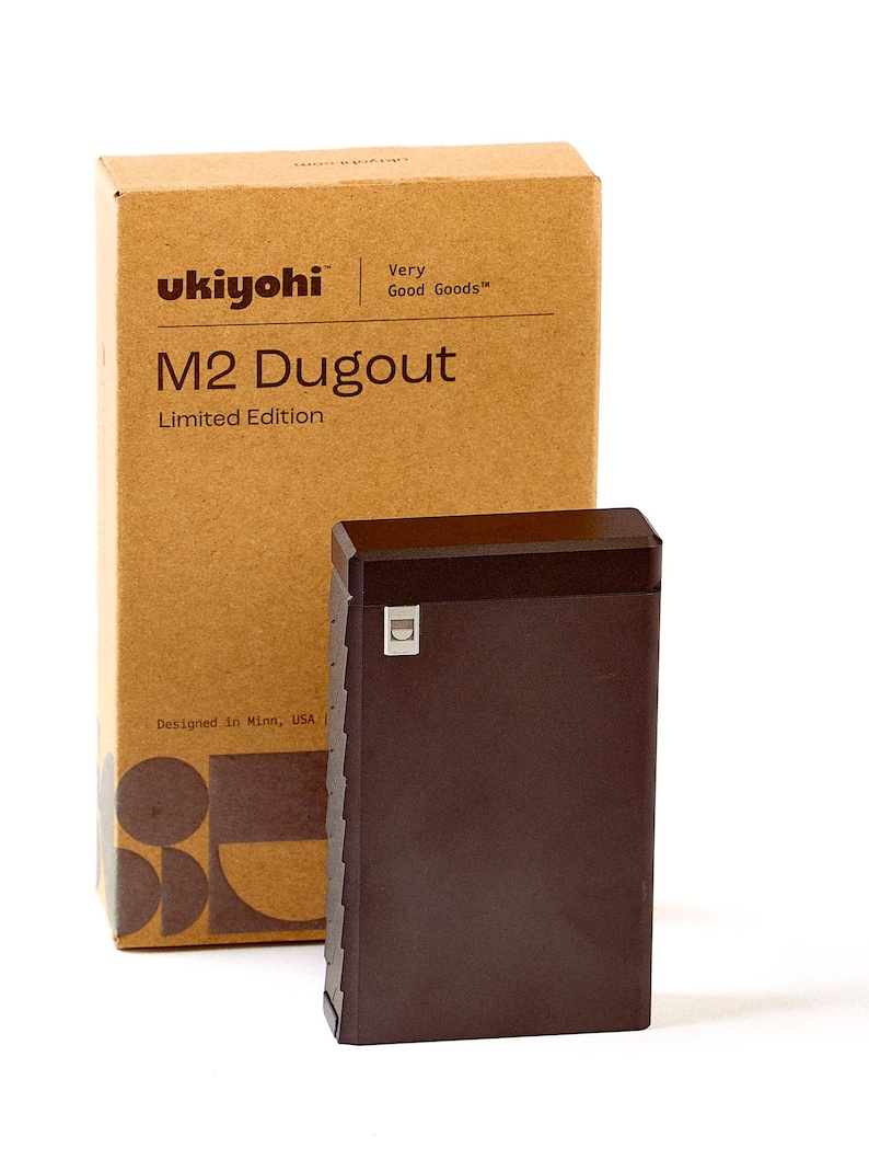 May include: A brown, rectangular box with the text "ukiyohi" and "M2 Dugout Limited Edition" printed on the front. The box is sitting on a white surface next to a brown, rectangular, metal container with a small, silver, rectangular logo on the front.