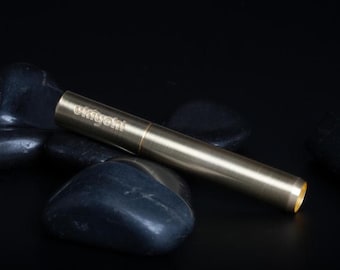 Könnte beinhalten: Ein goldfarbenes, zylindrisches Objekt mit dem Wort "ukiyoho" auf der Seite eingraviert. Das Objekt liegt auf einem Bett aus dunklen, glatten Steinen vor einem schwarzen Hintergrund. Das Objekt hat eine kleine Öffnung an einem Ende.