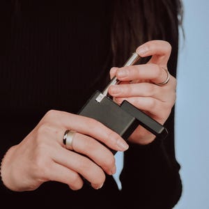 May include: A person holding a black and silver device with a silver metal rod extending from the top. The device has a rectangular shape and a small, square button on the side.