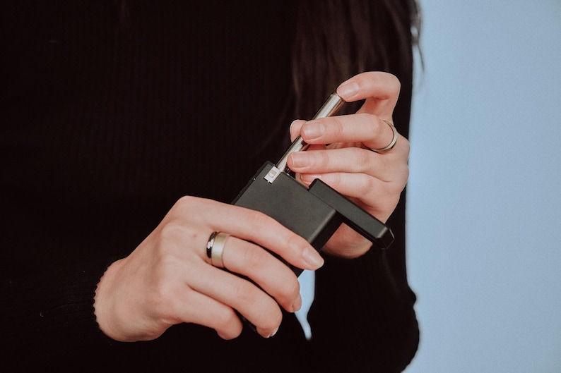 May include: A person holding a black and silver device with a silver metal rod extending from the top. The device has a rectangular shape and a small, square button on the side.