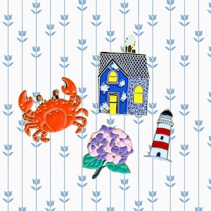 May include: A set of four enamel pins featuring a blue house, a red crab, a purple flower, and a red and white lighthouse. The pins are on a white background with a blue and white striped pattern.