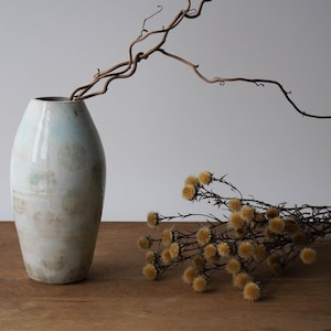 Könnte beinhalten: Eine hohe, cremefarbene Keramikvase mit hellblauem Anstrich, in der getrocknete Zweige stecken. Ein Bündel gelber, getrockneter Blumen liegt auf einer Holzoberfläche und erzeugt eine natürliche, minimalistische Ästhetik. Die Vase ist etwa 30 cm hoch.