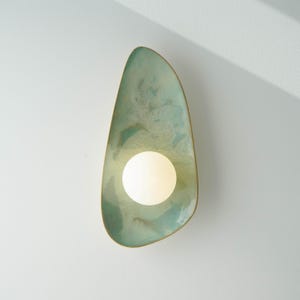 Puede incluir: Aplique de pared moderno con una forma orgánica única. La lámpara presenta una superficie texturizada de color verde claro con un borde dorado. Una bombilla redonda y blanca está centrada dentro del aplique, proporcionando una iluminación suave. El aplique está montado en una pared blanca.