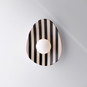 Peut inclure: Une applique murale composée d'une plaque arrière de forme ovale ornée de rayures verticales noires et crème. Une ampoule sphérique en verre blanc opaque est centrée sur la surface rayée, diffusant une lumière douce et uniforme.