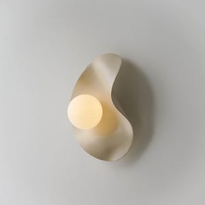 Könnte beinhalten: Cremefarbene Wandleuchte mit einer einzigartigen, organischen Form. Die Leuchte hat ein glattes, fließendes Design mit einer kugelförmigen, warm getönten Lichtquelle. Die Leuchte ist an einer neutralfarbenen Wand montiert.