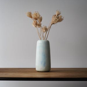 May include: A tall, light blue ceramic vase with a subtle ombre effect, holding dried, beige-colored ornamental grasses. The vase has a rounded base and a smooth, glossy finish, sitting on a dark wooden surface.