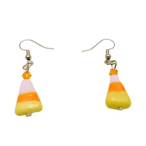 May include: A pair of candy corn earrings. Each earring features a silver-tone fish hook and a candy corn charm. The charms are white, orange, and yellow, with a small orange bead above the candy corn.