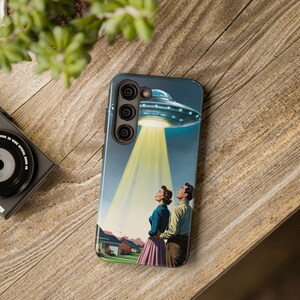Peut inclure: Une coque de téléphone avec une illustration rétro d'un OVNI projetant un faisceau jaune sur un couple. L'étui a un fond bleu avec un design vintage, et le couple regarde vers le haut l'engin spatial.