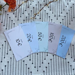 May include: Five pastel-colored gift certificates, each displaying a different Euro value: 10€, 20€, 50€, 100€, and 200€. The certificates have a decorative border and a sun design. The background is a white fabric with a black dotted pattern.