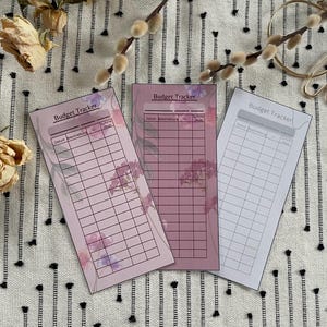 May include: Three budget tracker cards with floral designs. The cards are rectangular, with a pink grid pattern and the words "Budget Tracker" at the top. The cards have a floral design in shades of pink and purple.