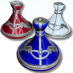 May include: Three tagine pots in red, blue, and white. Each pot has a conical lid and a round base. The pots are decorated with silver-coloured metal filigree and trim. These tagines are likely used for cooking traditional dishes, with a diameter of approximately 20 cm.