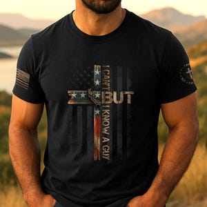 Pode incluir: T-shirt preto com um design de cruz que incorpora a bandeira americana e o texto "I CAN'T BUT I KNOW A GUY". A camisa tem gola redonda e mangas curtas. O design inclui estrelas e listras.