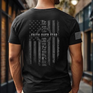 May include: Black t-shirt featuring a distressed American flag design with the words "Faith Over Fear" and a cross graphic. The back of the shirt also includes a quote from Psalm 118:6. A small American flag is on the sleeve.