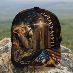 May include: A baseball cap featuring a lion, a cross, and an American flag design. The word "REDEEMED" is printed vertically on the right side of the cap. The text "YOUR TEXT" is printed on the bottom left side of the cap.
