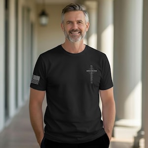 May include: Black t-shirt with a small American flag on the left sleeve. The shirt features a cross design with the words "FAITH OVER FEAR" and "INSPIRING" printed vertically. The person is smiling and standing in front of a white columned structure.