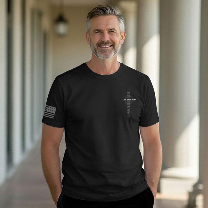 May include: Black t-shirt with a small American flag on the left sleeve. The shirt features a cross design with the words "FAITH OVER FEAR" and "INSPIRING" printed vertically. The person is smiling and standing in front of a white columned structure.