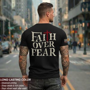 May include: Black t-shirt with the words "FAITH OVER FEAR" in distressed white font and a red cross. The shirt has a crew neck and short sleeves. The image also shows the back of a person with tattoos on their arms.