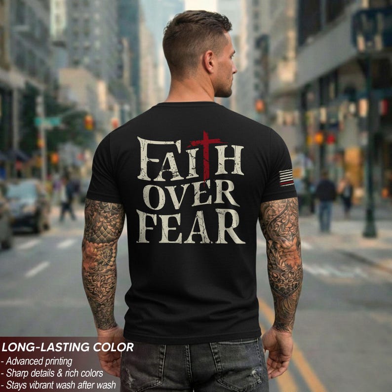 May include: Black t-shirt with the words "FAITH OVER FEAR" in distressed white font and a red cross. The shirt has a crew neck and short sleeves. The image also shows the back of a person with tattoos on their arms.