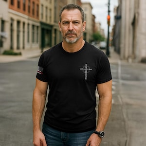 May include: Black crew neck t-shirt with a cross graphic and the text "FAITH OVER FEAR". The shirt also features a small American flag design on the left sleeve. The person is wearing blue jeans and a black watch.