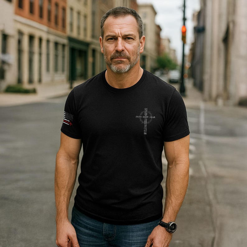 May include: Black crew neck t-shirt with a cross graphic and the text "FAITH OVER FEAR". The shirt also features a small American flag design on the left sleeve. The person is wearing blue jeans and a black watch.