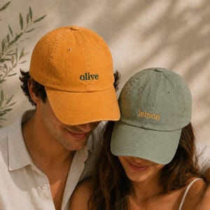 Op de afbeelding: Twee baseballpetten, één mosterdgeel en één saliegroen. Op de gele pet staat het woord "olive" in zwart geborduurd, en op de groene pet staat "lemon" in oranje geborduurd. De petten worden door twee personen gedragen.